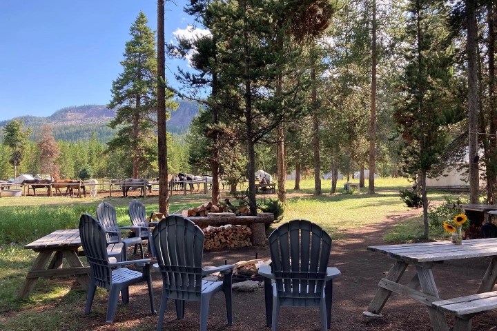 Outdoor picnic area with chairs around a fire pit and horses in a fenced area nearby.