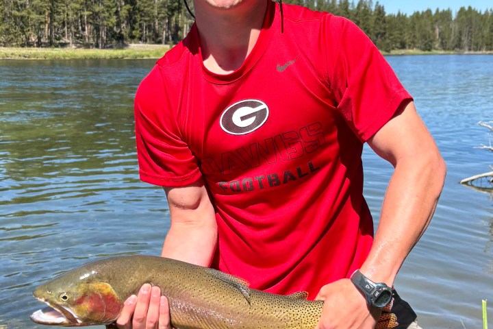 Person in red shirt holds a large fish by a lake, wearing an orange cap and sunglasses.