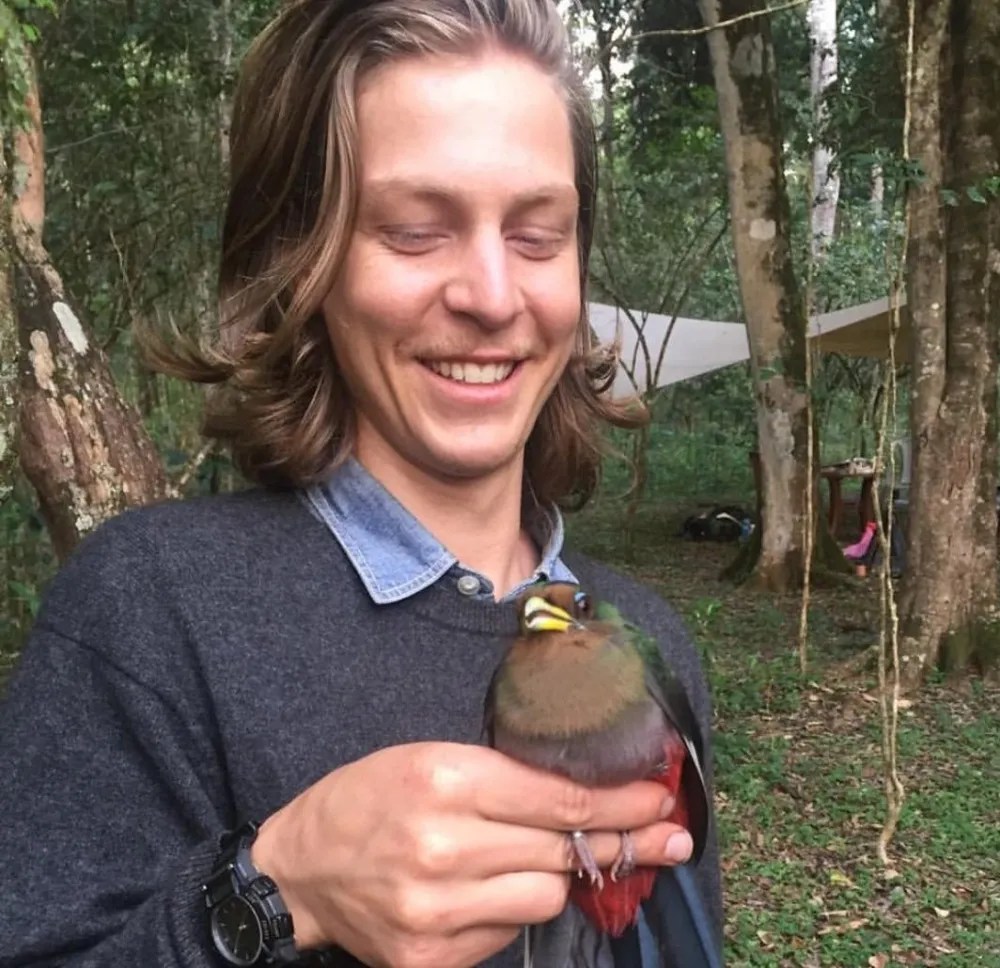 Person smiling, holding a colorful bird in a forest setting.
