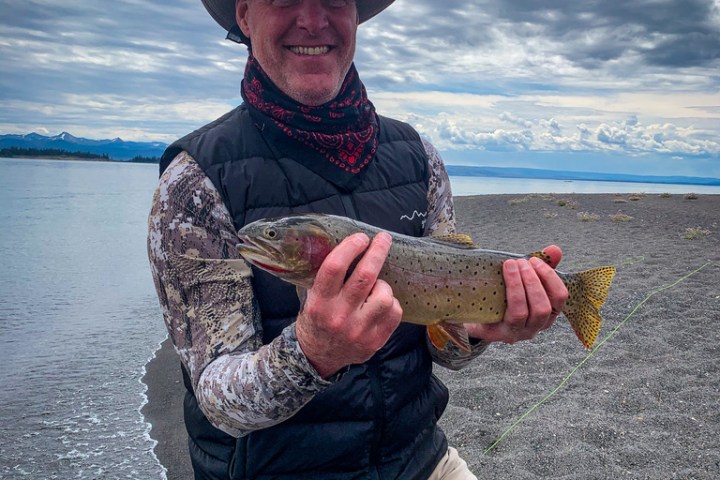 Person in a hat holding a fish by a lake under a cloudy sky.