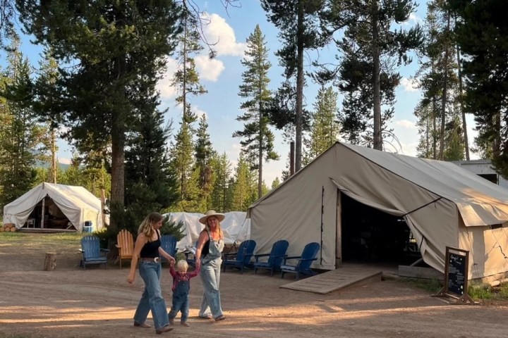 Family walking near canvas tents in a forest campground with blue sky.