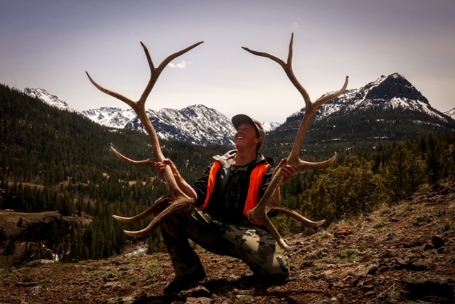 Person holding antlers in a mountainous landscape with snow-capped peaks in the background.